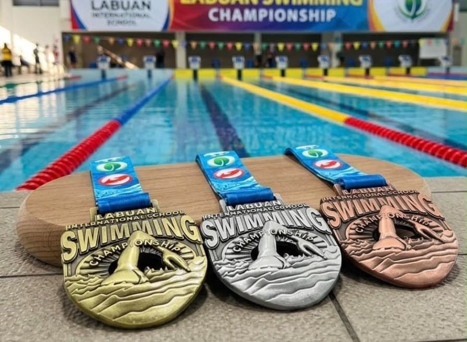 Gold, silver and bronze Labuan International School Swimming Championship medals on a wooden stand beside the pool